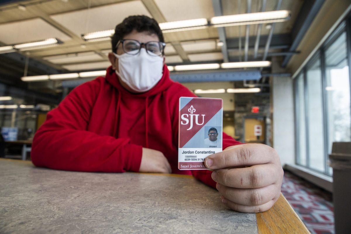 Constantino is wearing a red hoodie and a white mask and is holding his ID up in the foreground. He is in Post Learning Commons, sitting at a table.