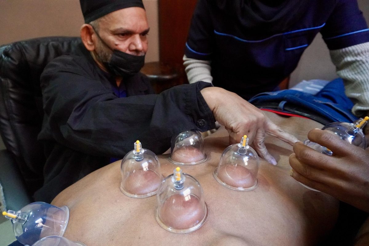Malik Cassim, using what his wife Sadia Cassim calls the “gift in his hands,” presses on a patient’s back to show his assistants where cups should be placed. Cassim lost his ability to speak following a stroke in 2022. PHOTO: CARLINA HERSHOCK/THE HAWK