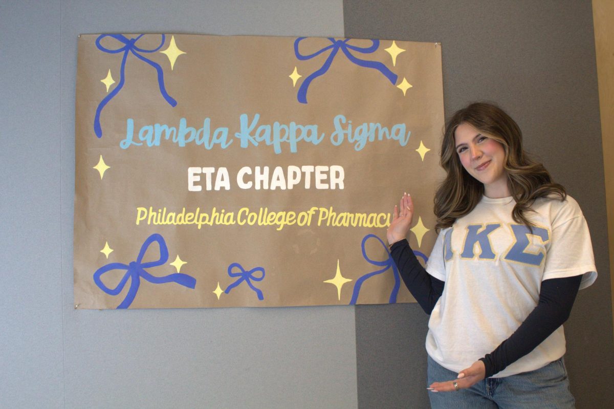 Caterina Pica ’25, PharmD ’27 poses with a banner for the Eta chapter of Lambda Kappa Sigma, an organization dedicated to “empowering women in pharmacy.” 
PHOTO: PRESTON THOMAS '28/THE HAWK
